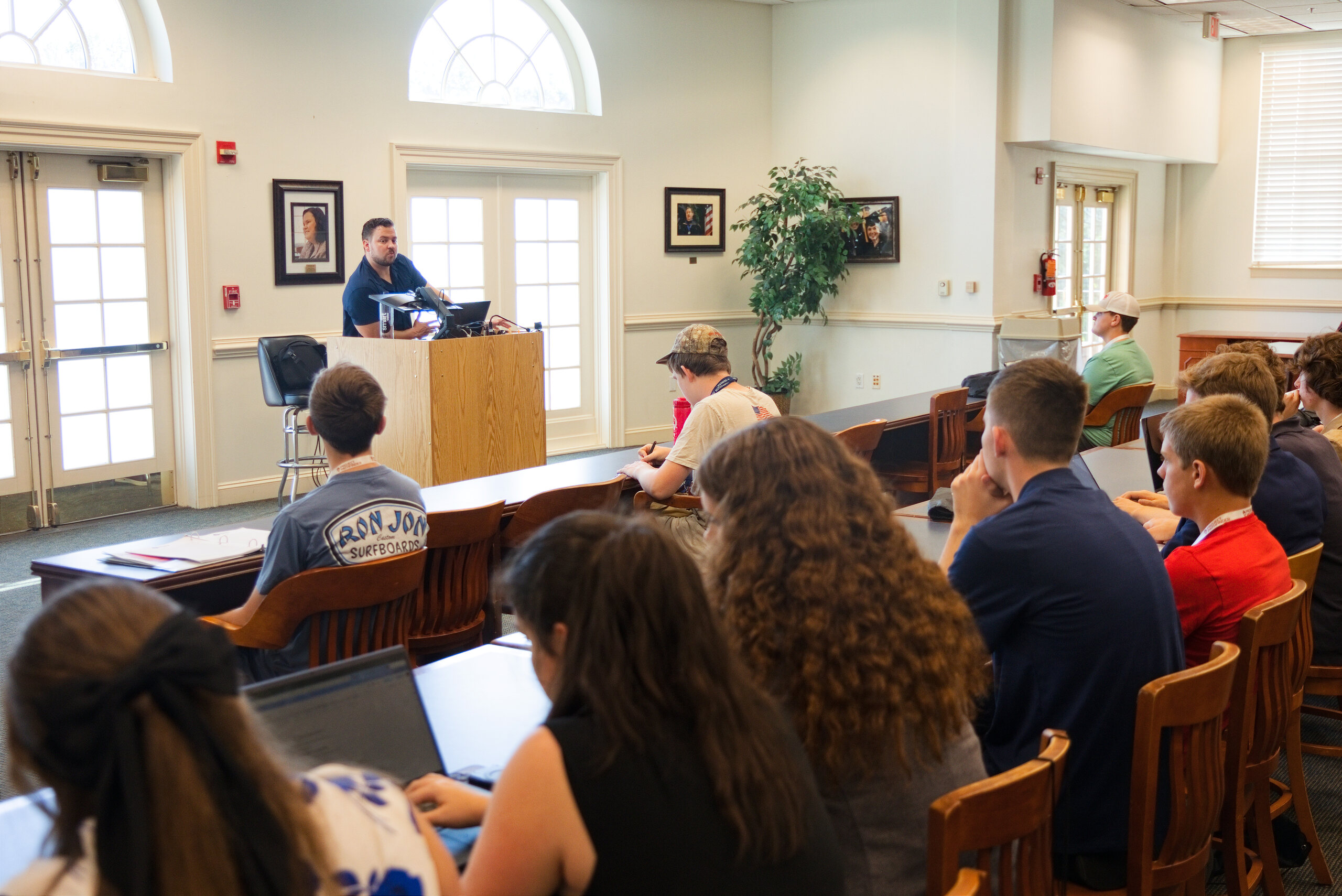 Speaker at a wooden podium addresses students seated at long tables in a bright hall