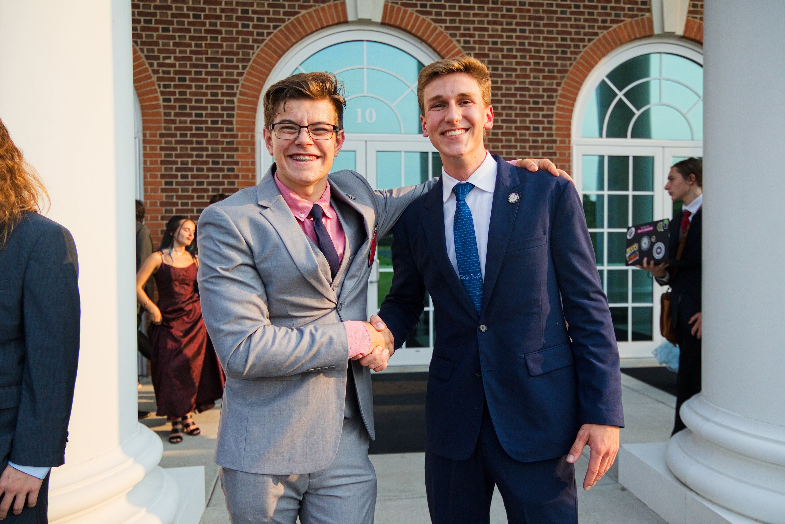 Two smiling students shake hands beneath arched windows at a brick building entrance